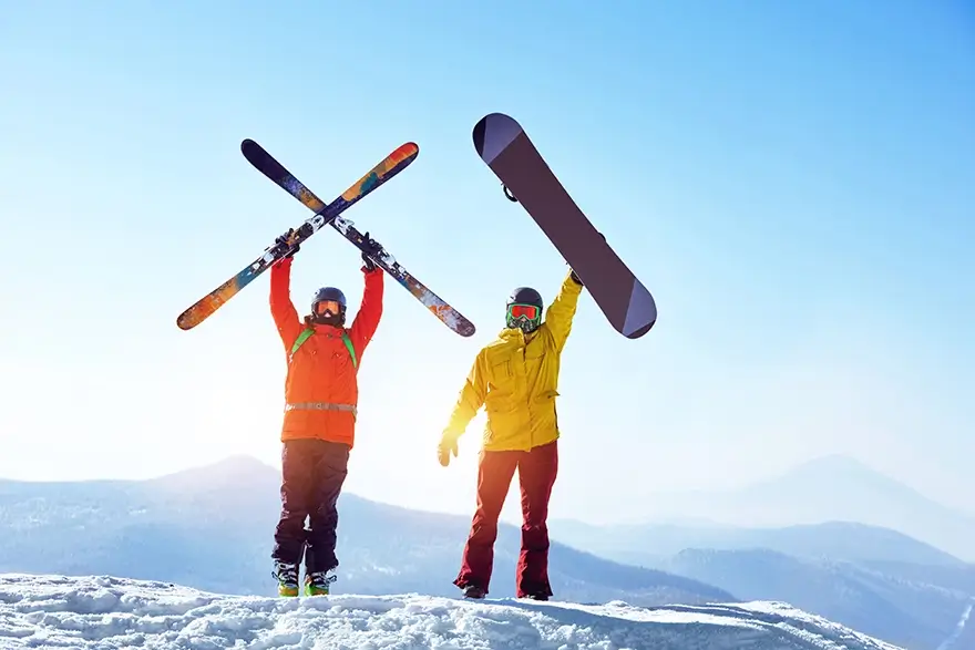Snowboarder and skier holding their snow equipment above their heads in triumphant on top of the Snowy mountains.