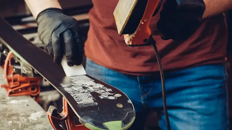 Skis being waxed in a workshop by an employee wearing safety gloves.